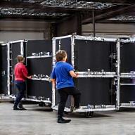 Miembros del equipo de stage hand trabajando en conjunto para montar rápidamente el equipo en un escenario, con cajas de transporte al fondo.